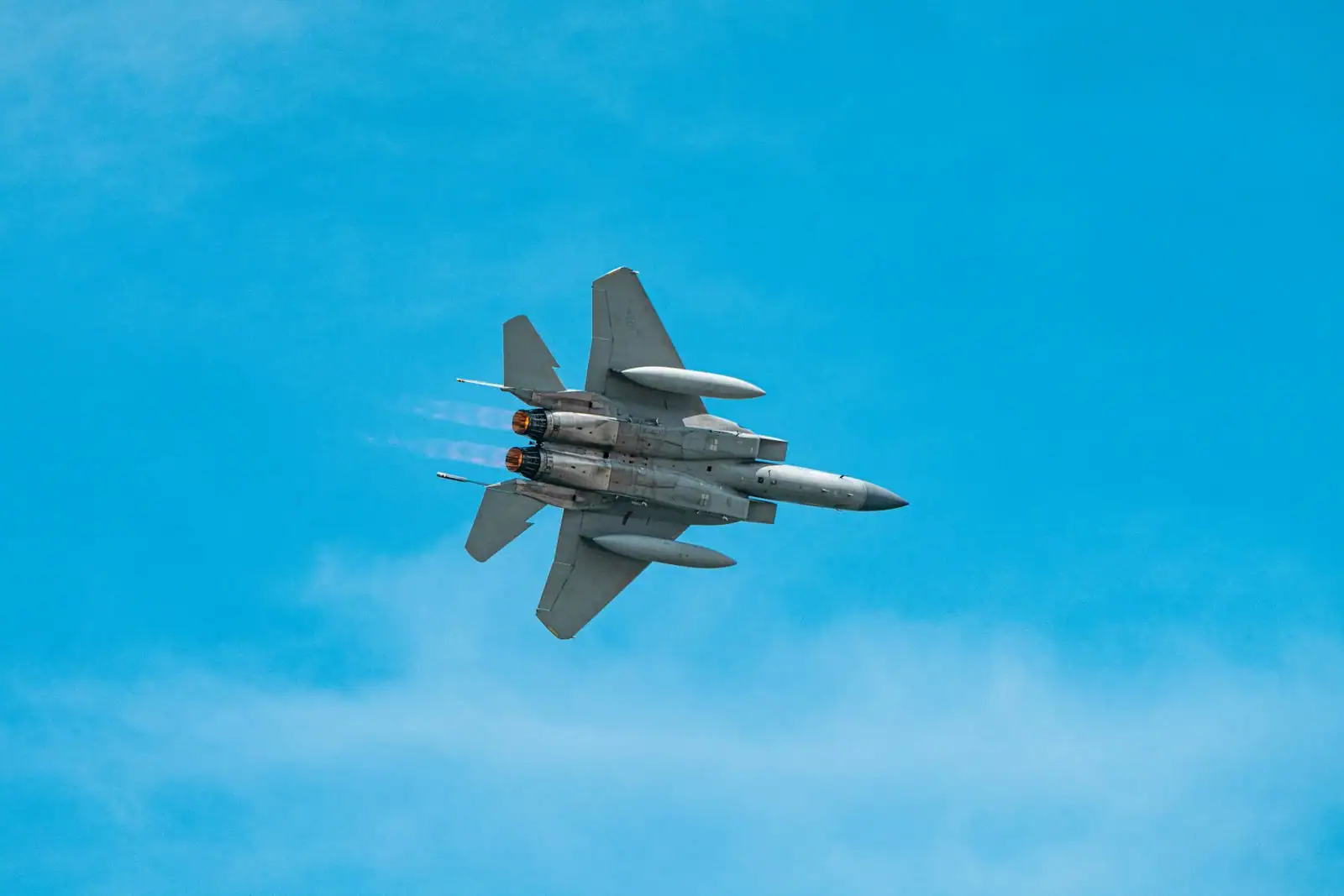 Avion de chasse militaire à réaction en vol dans un ciel bleu dégagé, vu de dessous, réacteurs allumés.