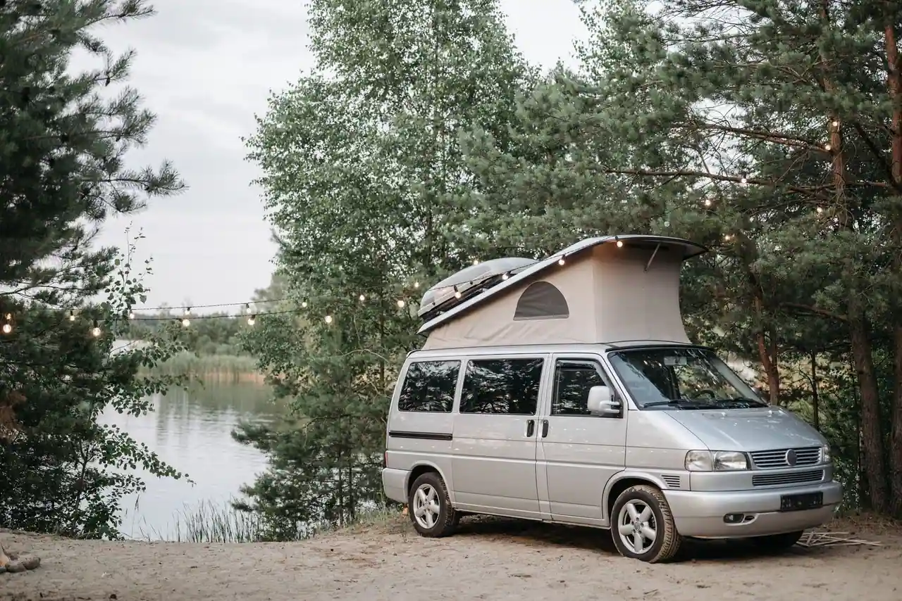 Van aménagé avec tente de toit déployée, stationné au bord d'un lac bordé d'arbres dans un camping.
