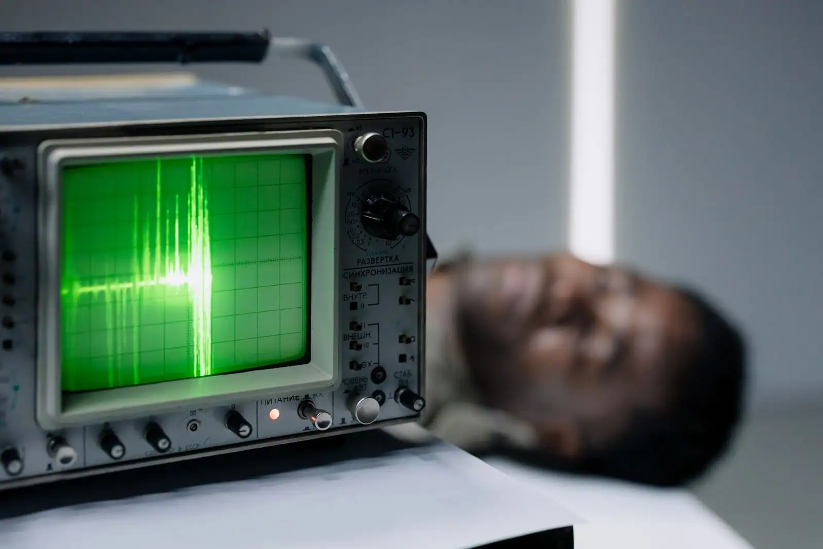 Oscilloscope analogique affichant une trace verte de signal sur son écran, une personne floue en arrière-plan