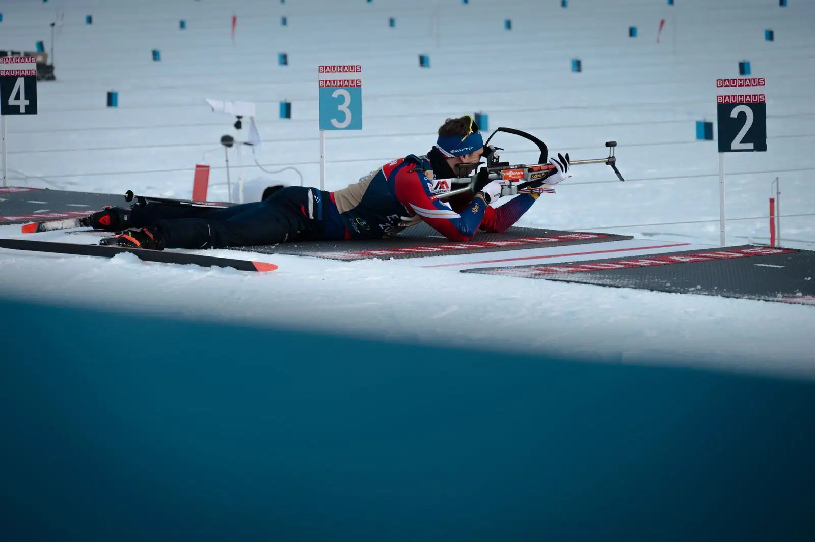 Biathlète allongé au pas de tir, en position de tir avec sa carabine, sur un stade de biathlon enneigé