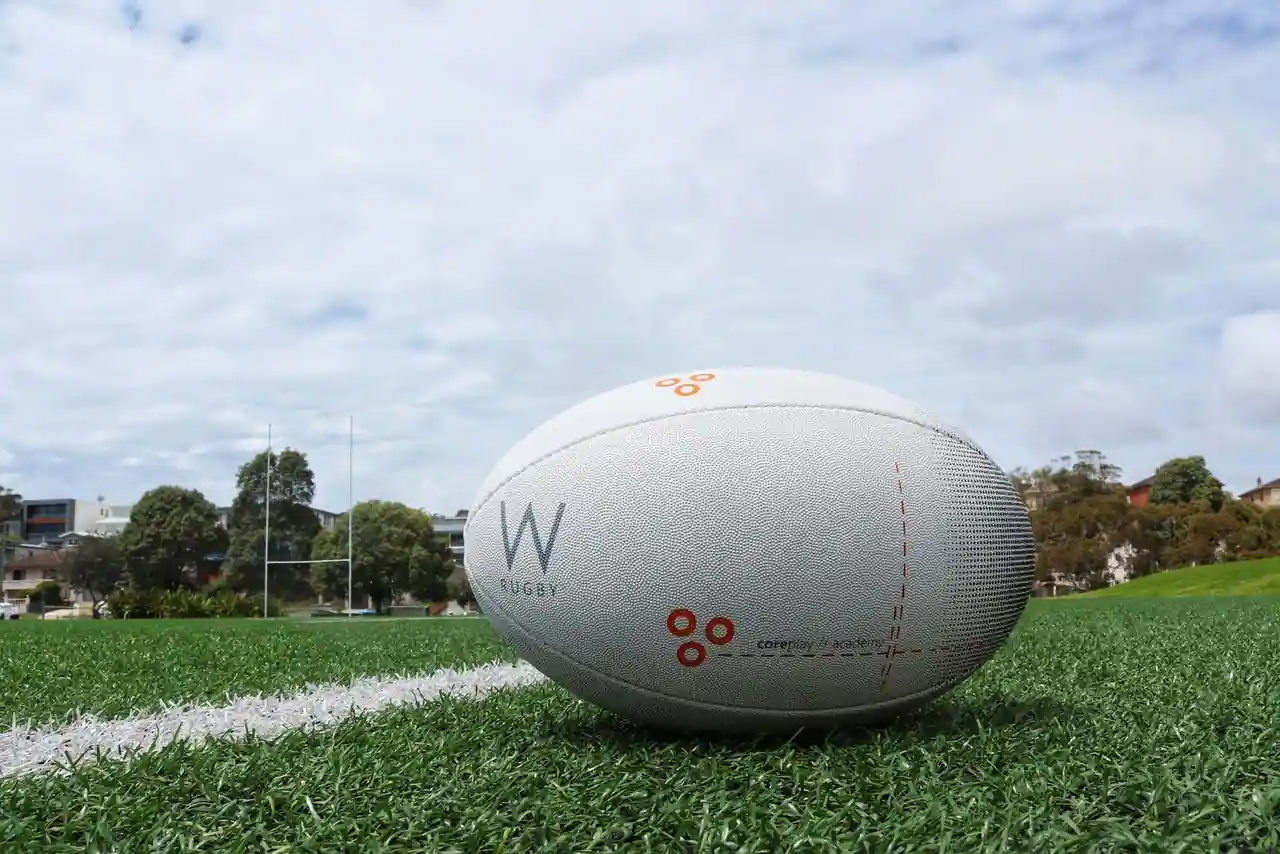 Ballon de rugby blanc posé sur la pelouse verte d'un terrain, poteaux de rugby en arrière-plan