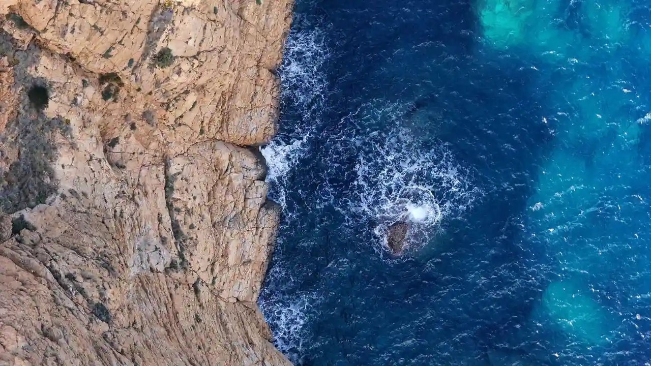 Vue aérienne d'une côte méditerranéenne rocheuse aux eaux turquoise, ourlée d'écume blanche.