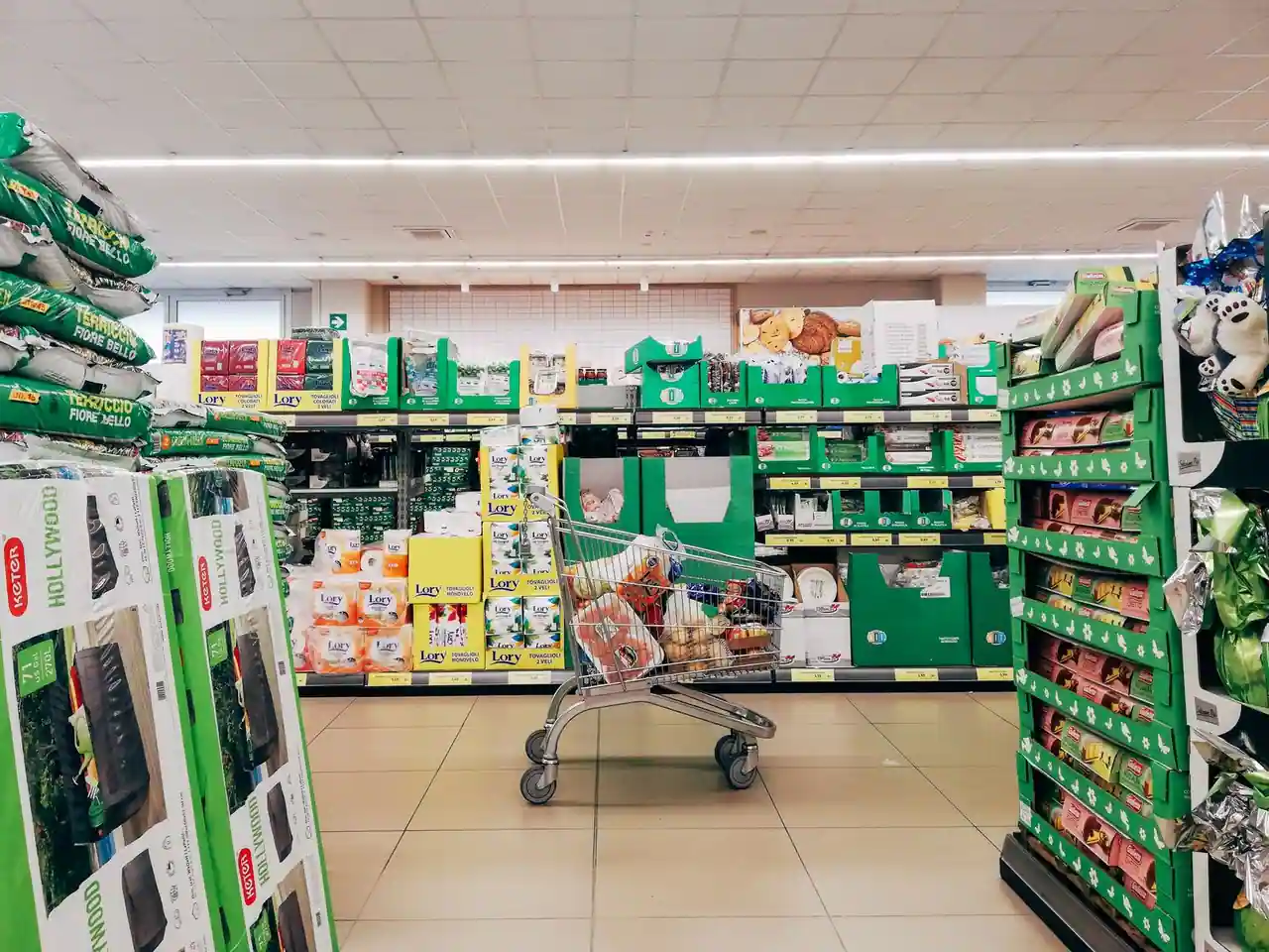 Caddie rempli de courses dans une allée de supermarché, devant des rayons et des étiquettes de prix