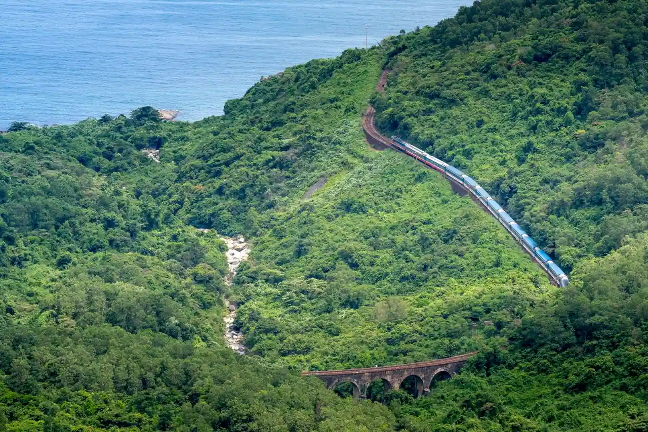 Train de voyageurs serpentant à flanc de montagne boisée au-dessus de la mer, près d'un viaduc.