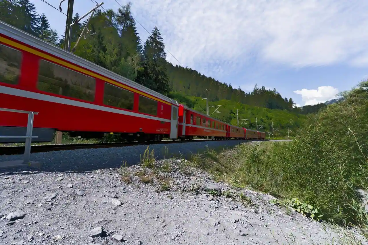 Train rouge de montagne longeant un versant boisé verdoyant par beau temps, image illustrative.