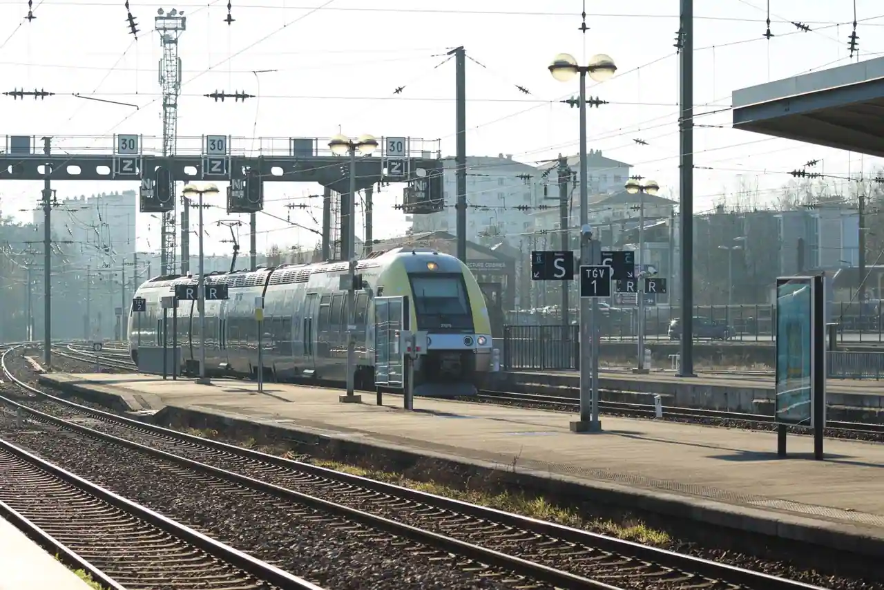 Train régional à quai dans une gare, voies et caténaires sous le soleil.