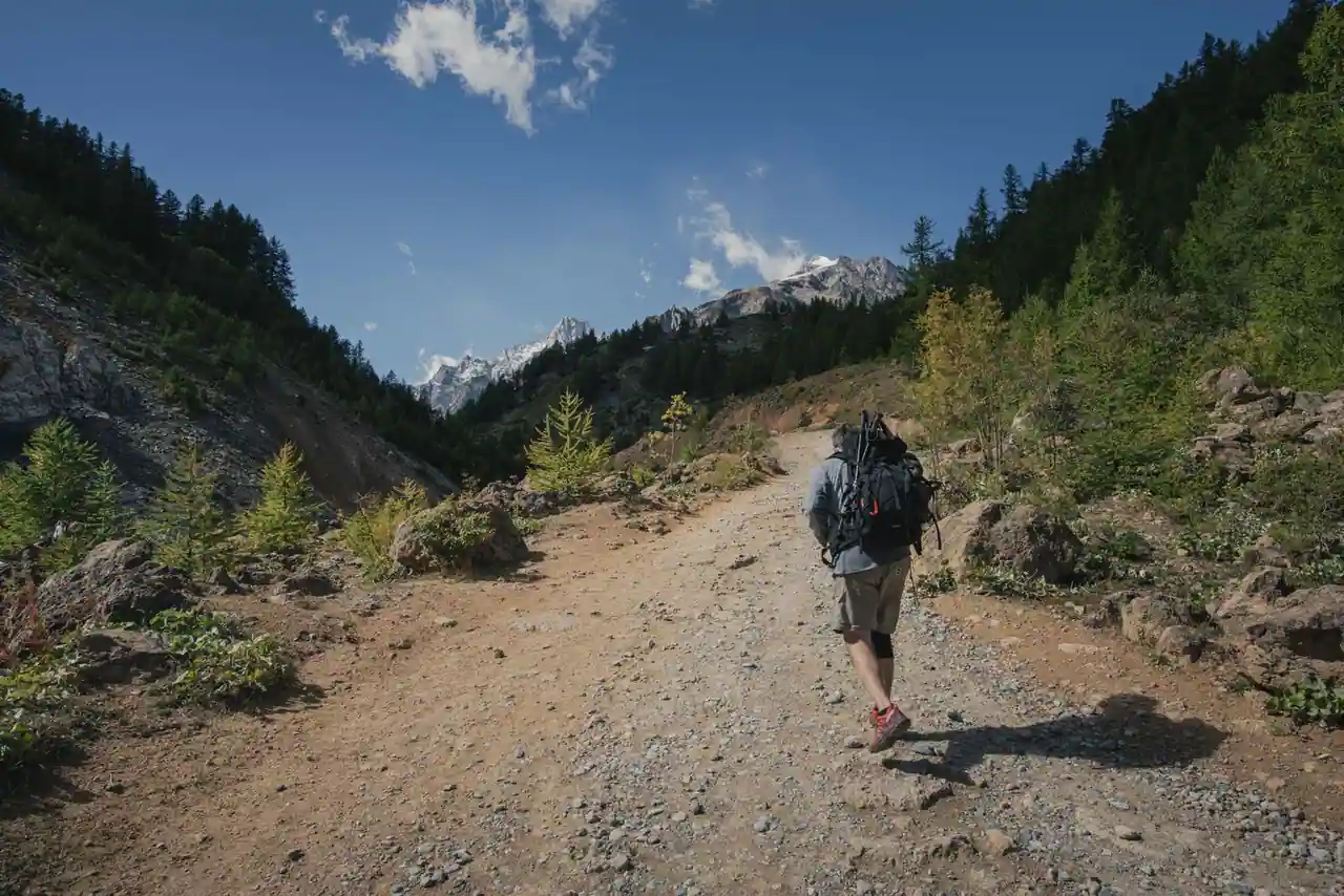 Un randonneur portant un grand sac à dos marche sur un chemin de terre en montagne, sommets en arrière-plan.