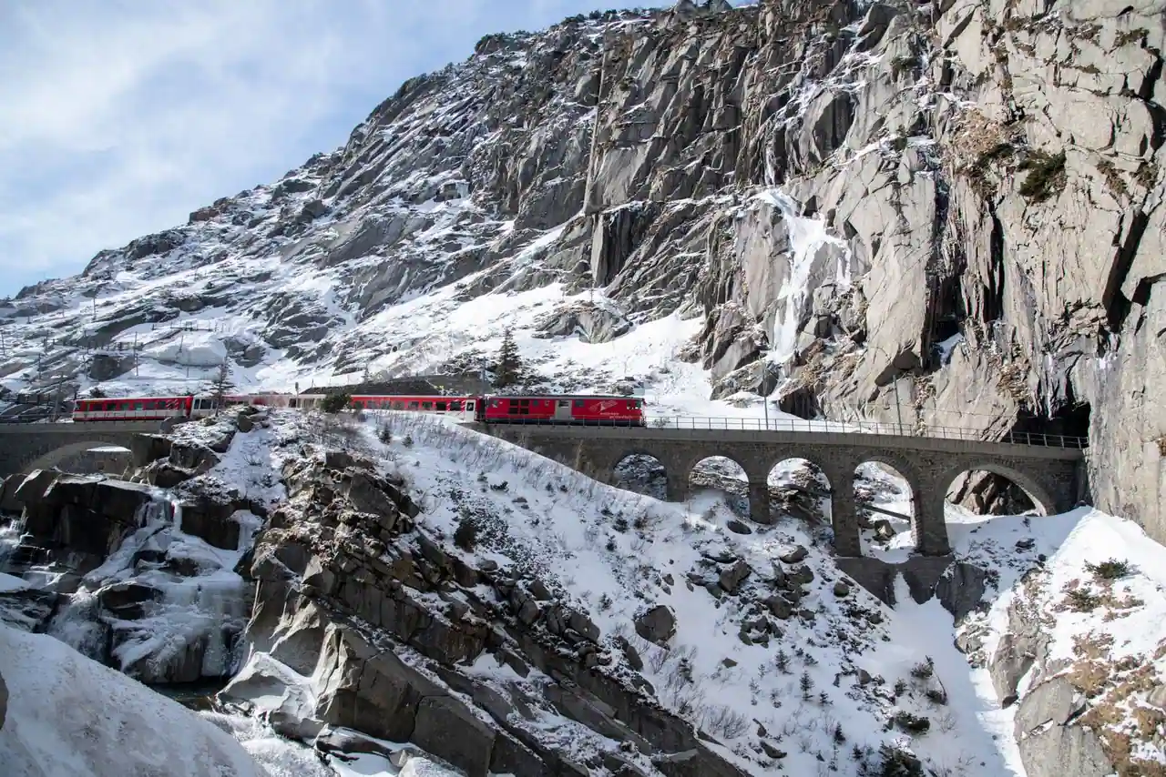 Train rouge longeant un paysage de montagnes suisses, viaduc et sommets enneigés en arrière-plan