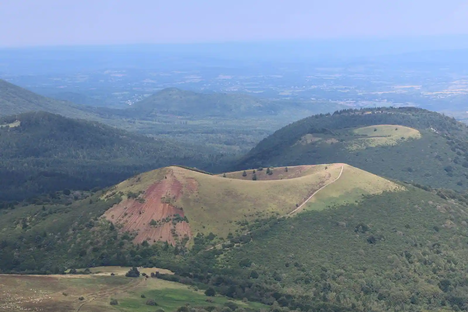 Cratère volcanique herbeux de la chaîne des Puys, coulée de scories rouges et plaine en arrière-plan.