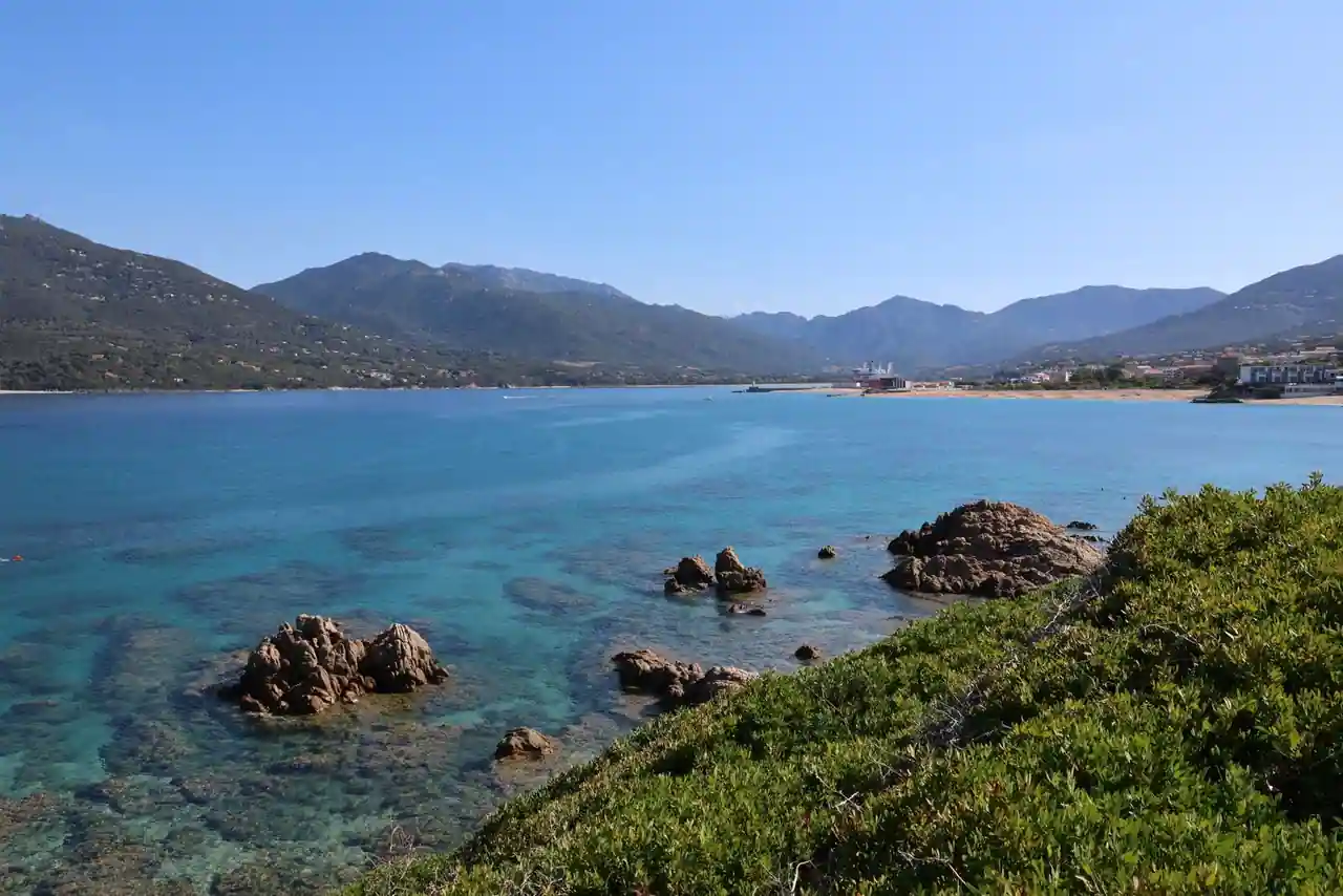 Un golfe corse aux eaux claires, rochers affleurants et maquis au premier plan, montagnes au fond.
