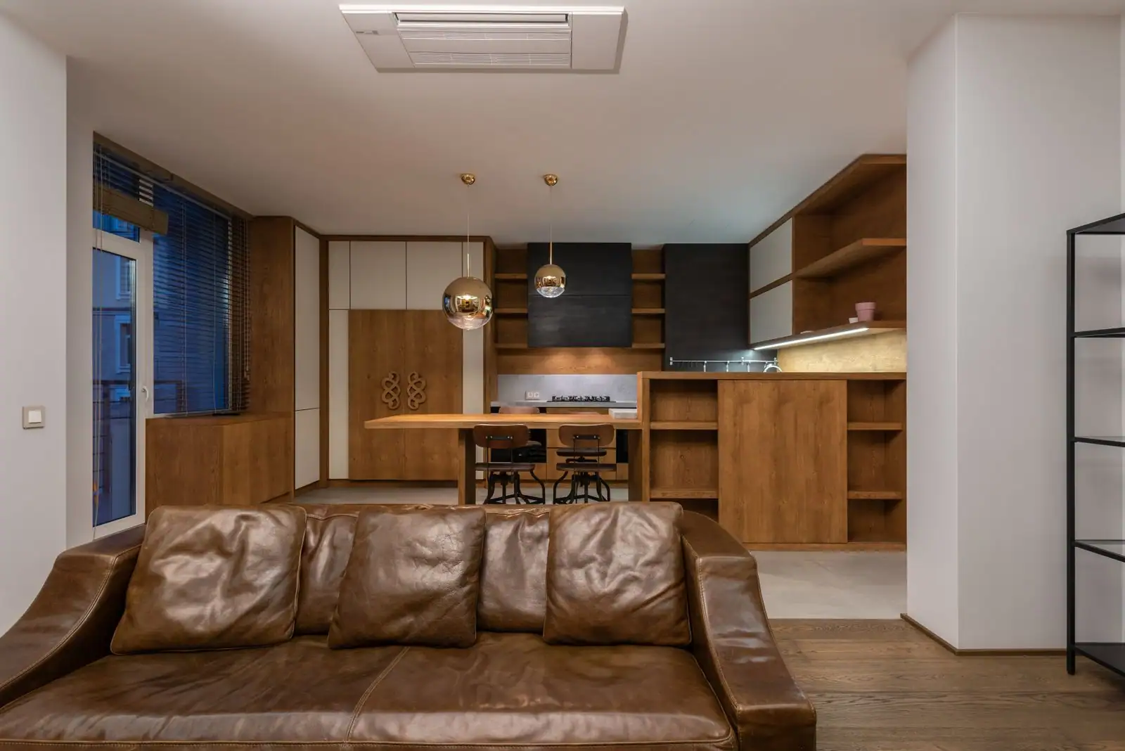 Séjour d'un appartement meublé, canapé en cuir au premier plan et cuisine ouverte en bois à l'arrière.