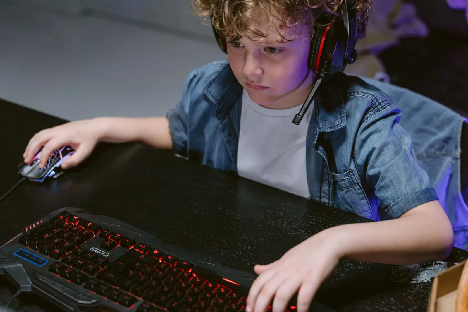 Un enfant concentré joue sur un ordinateur, une main sur la souris, l'autre sur un clavier rétroéclairé.