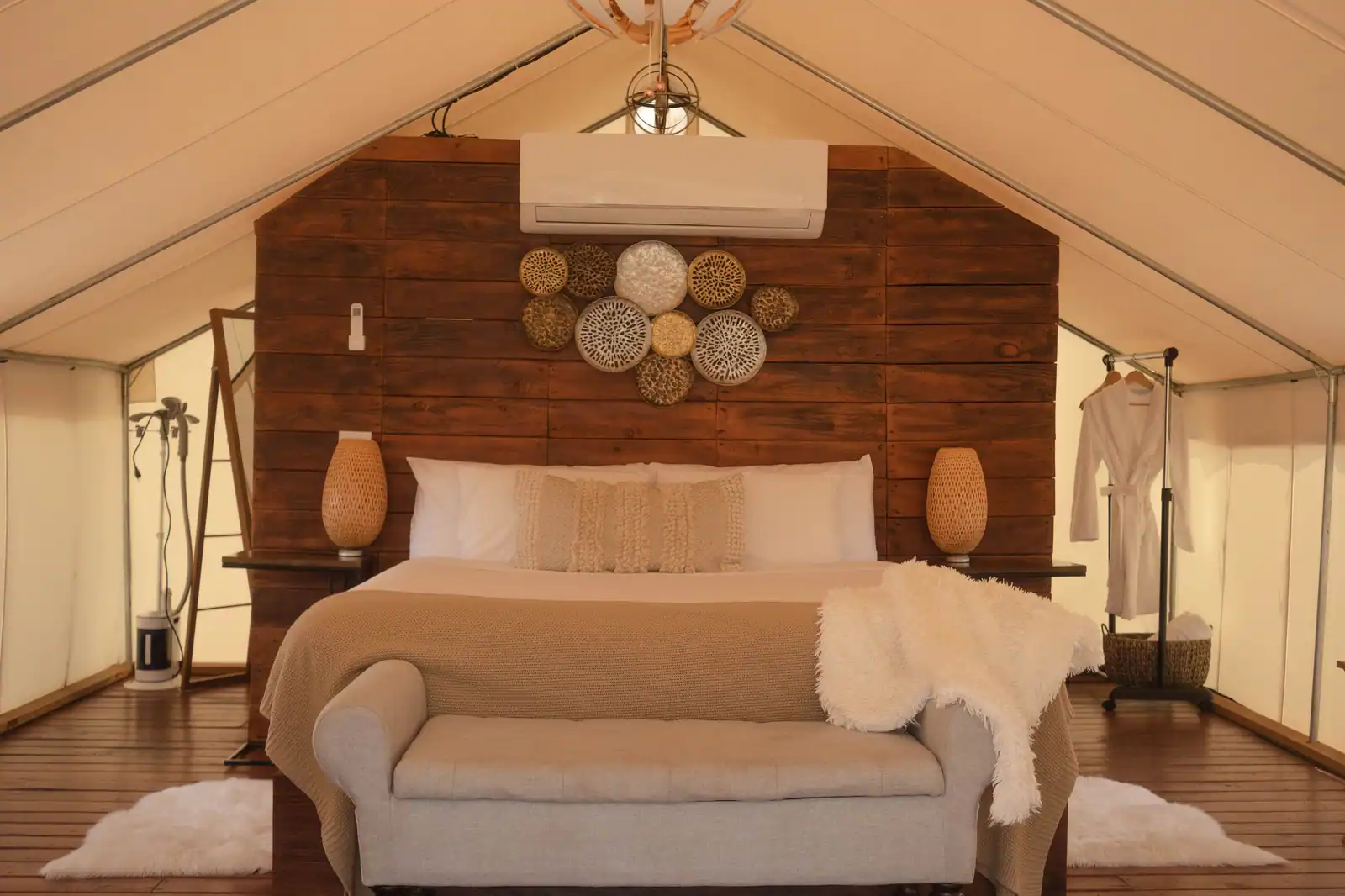 Intérieur d'une tente de glamping : lit fait sur plancher de bois, banquette et peignoirs suspendus.