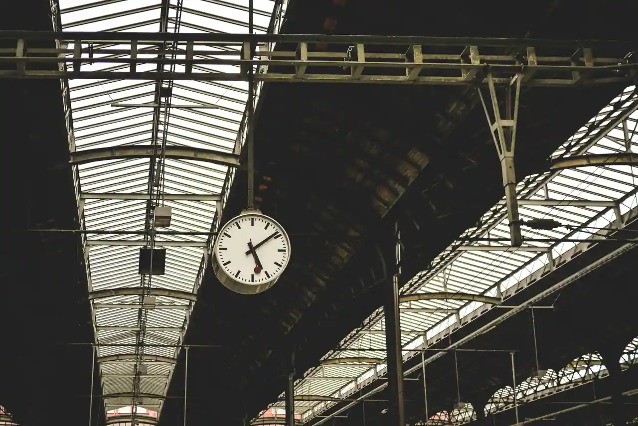 Horloge de gare suspendue sous la verrière d'un quai, cadran clair à aiguilles noires.