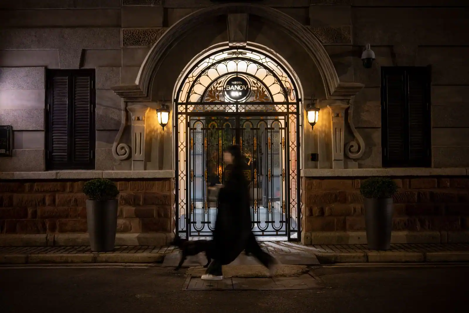 Un passant longe le trottoir devant l'entrée éclairée d'un hôtel, sous un porche en fer forgé, la nuit.