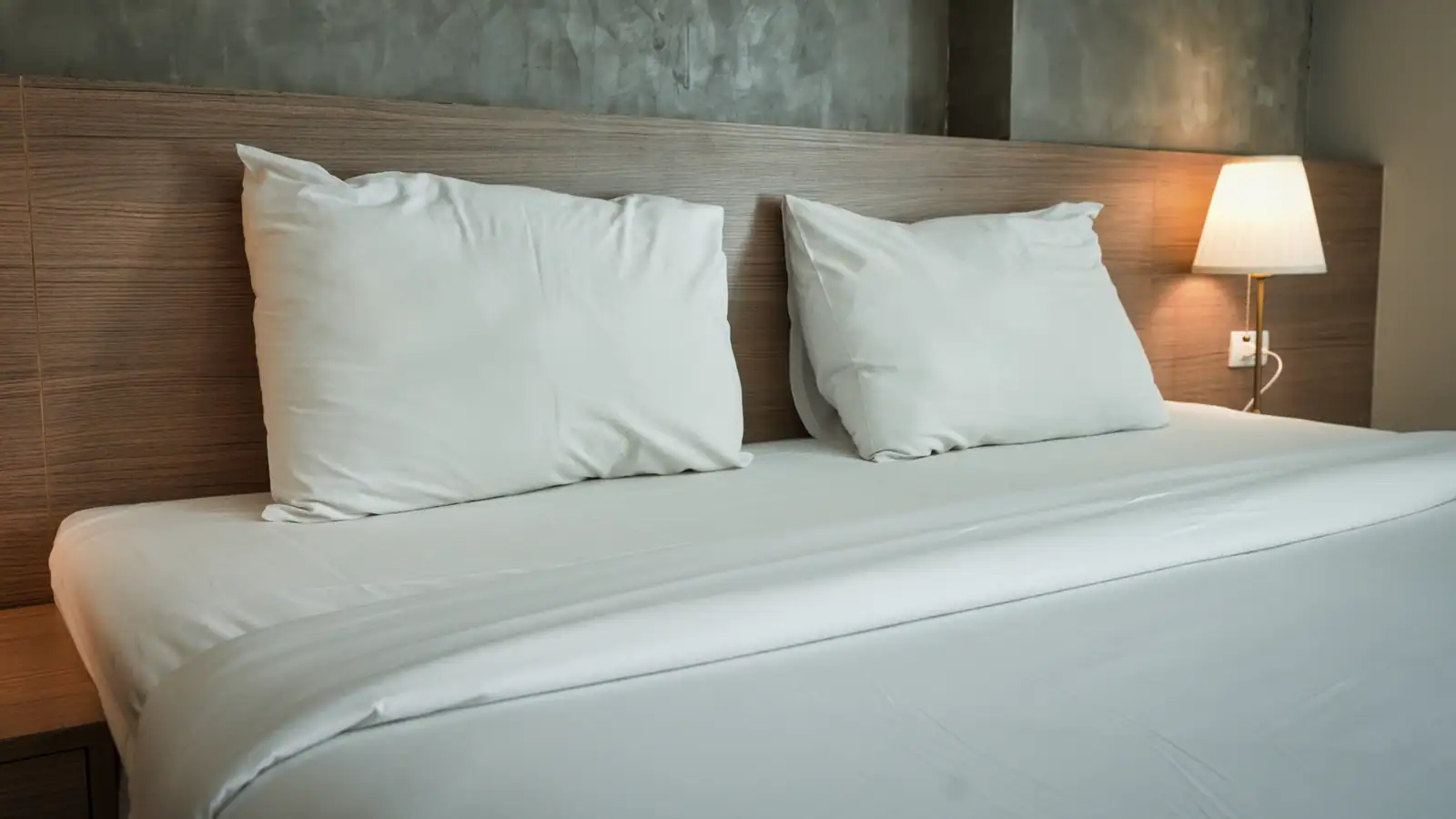 Lit fait dans une chambre d'hôtel sobre, deux oreillers blancs contre une tête de lit en bois et une lampe de chevet