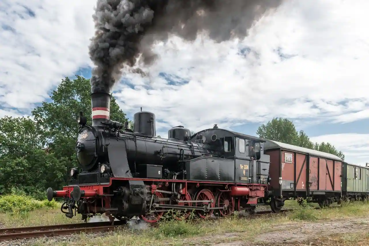 Locomotive à vapeur noire à roues rouges sous son panache de fumée, en tête d'un convoi ancien.