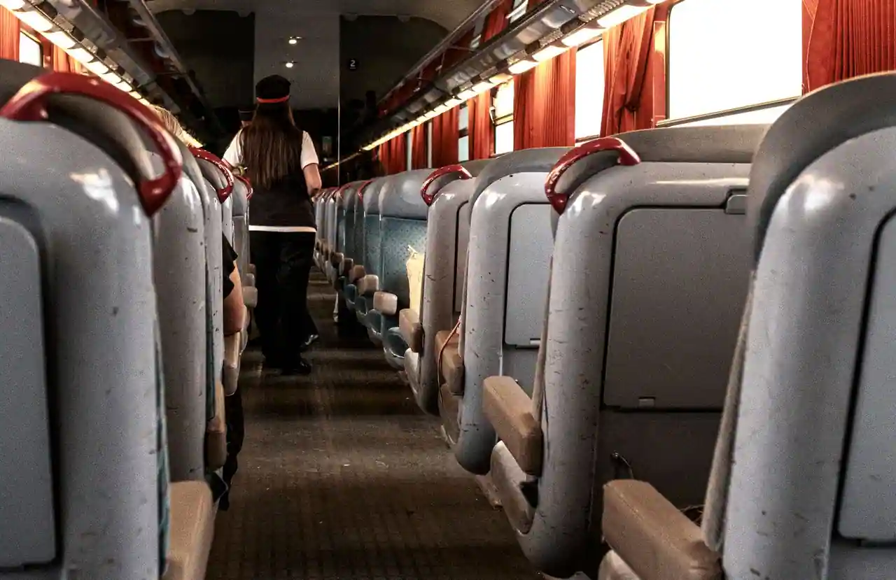 Couloir central d'une voiture de train ancienne, sièges gris à poignées rouges et rideaux aux fenêtres
