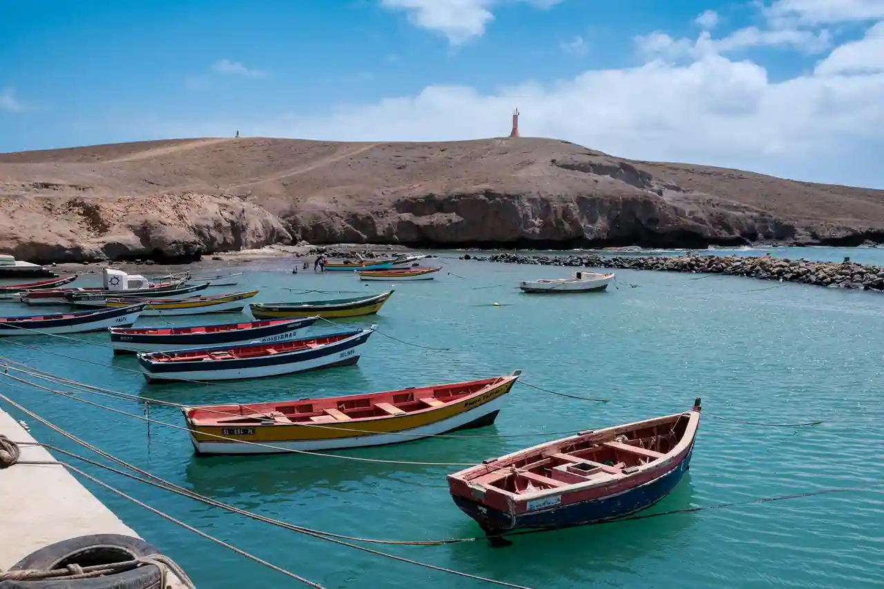 Des barques de pêche colorées mouillées dans une baie calme, au pied d'une colline aride du Cap-Vert.