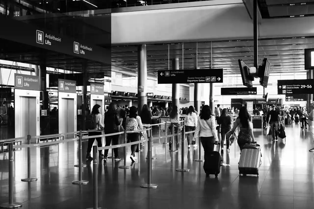 Voyageurs avec bagages se dirigeant vers les zones d'enregistrement d'un aéroport, en noir et blanc.