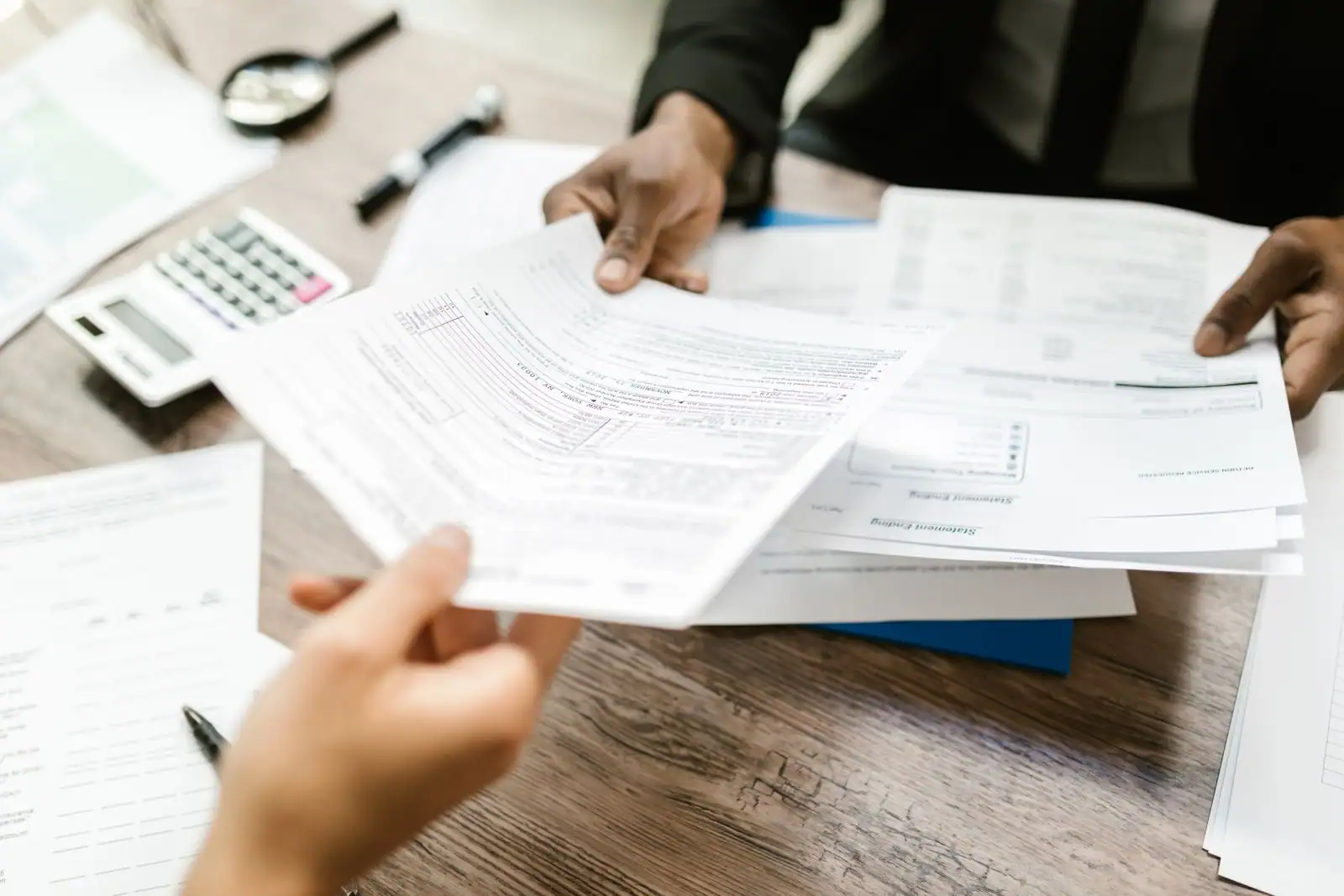 Deux personnes qui échangent des documents administratifs sur un bureau, dossiers et calculatrice à proximité.